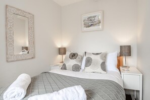 Un lit bien fait, recouvert d’une couette grise, avec des oreillers blancs ornés d’un motif de feuilles, un grand miroir doté d’un cadre décoratif et un tableau encadré accroché au mur.