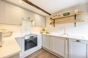 A kitchen with white cabinets, a built-in oven, a sink with a gold faucet, and wooden shelves above the counter. - 1 Bed in Frampton On Severn (oc-c32001) (Frampton on Severn)