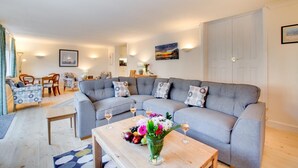 A modern living room with a grey sectional sofa, a wooden coffee table with fruit and flowers, and a dining area with a table and chairs.