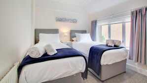 A double room with two beds, each with white linens and a blue blanket. There is a window with striped curtains, a radiator, and a wall-mounted artwork.