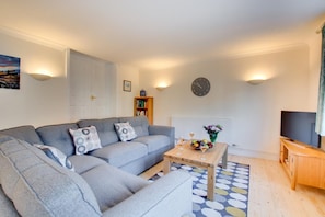 A living room with a grey sofa, a wooden coffee table with decorative items, a bookshelf, a clock, and a television on a wooden stand. - 4 Bed in Cromer (oc-2069) (Cromer)