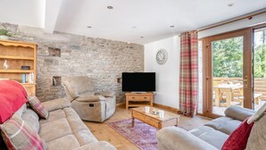 A cozy living room with a stone wall, a flat-screen TV, and a large window with curtains. - 3 Bed in Llangadog (oc-c32033) (Llangadog)