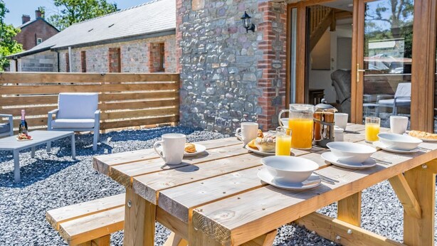 An outdoor dining area with a stone wall, wooden furniture, and a breakfast setup including plates, cups, and a pitcher of orange juice. - 3 Bed in Llangadog (oc-c32033) (Llangadog)