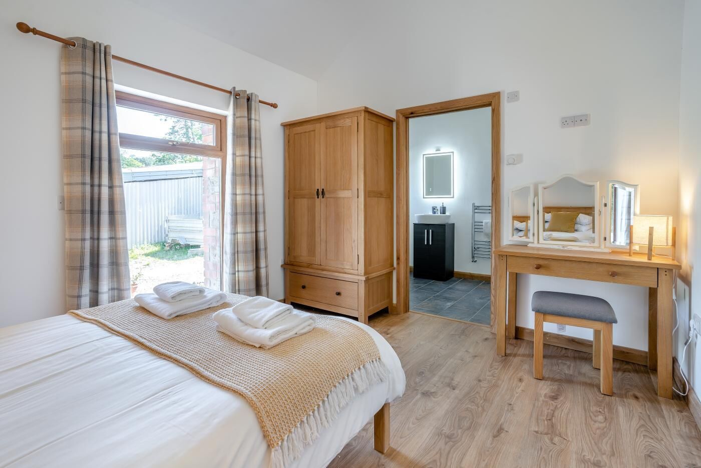 A bedroom with a bed, a wooden wardrobe, a desk with a mirror, and a bathroom visible through an open door.