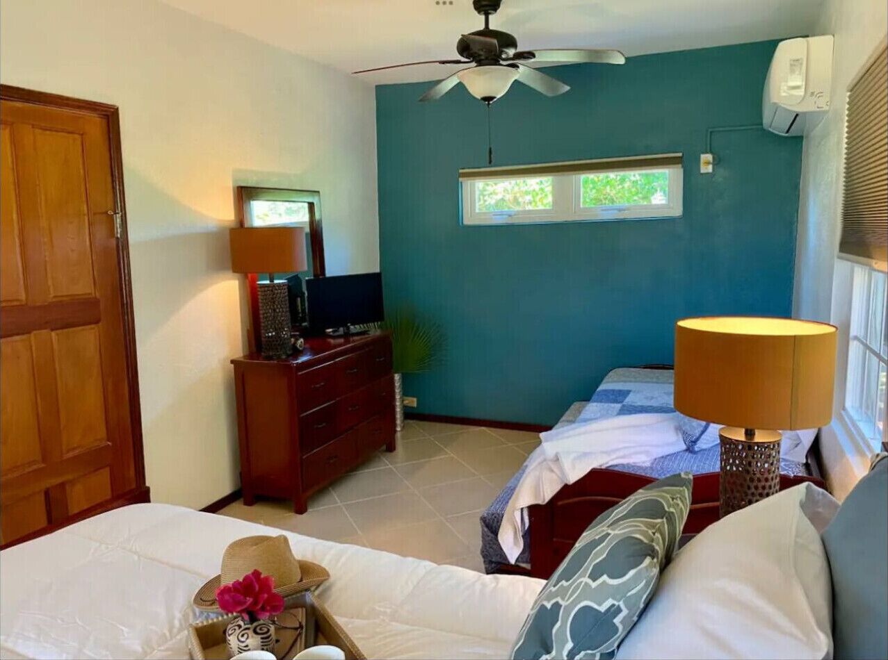 Une chambre avec deux lits, un ventilateur de plafond, un téléviseur posé sur une commode et une fenêtre avec des stores.