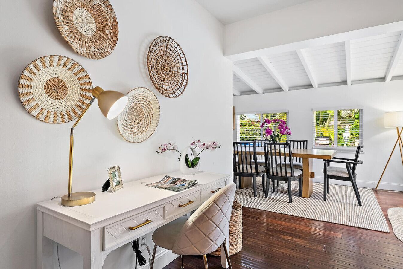 A modern dining area with a white table and chairs, a woven wall hanging, and a view of greenery outside.