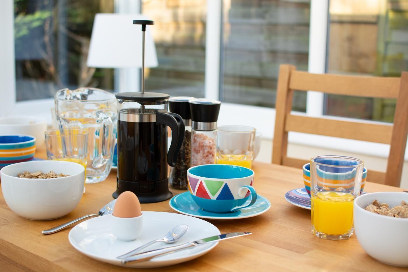 Una mesa de desayuno con una cafetera, un hervidor de té, una taza de café, un tazón de cereal, un vaso de jugo de naranja y un plato con un huevo duro.