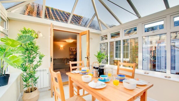Une véranda avec une table à manger dressée pour le petit-déjeuner, entourée de plantes en pot et de grandes fenêtres.