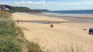 Una playa con una bandera roja que indica prohibido bañarse, algunas personas caminando y una sombrilla azul sobre la arena.