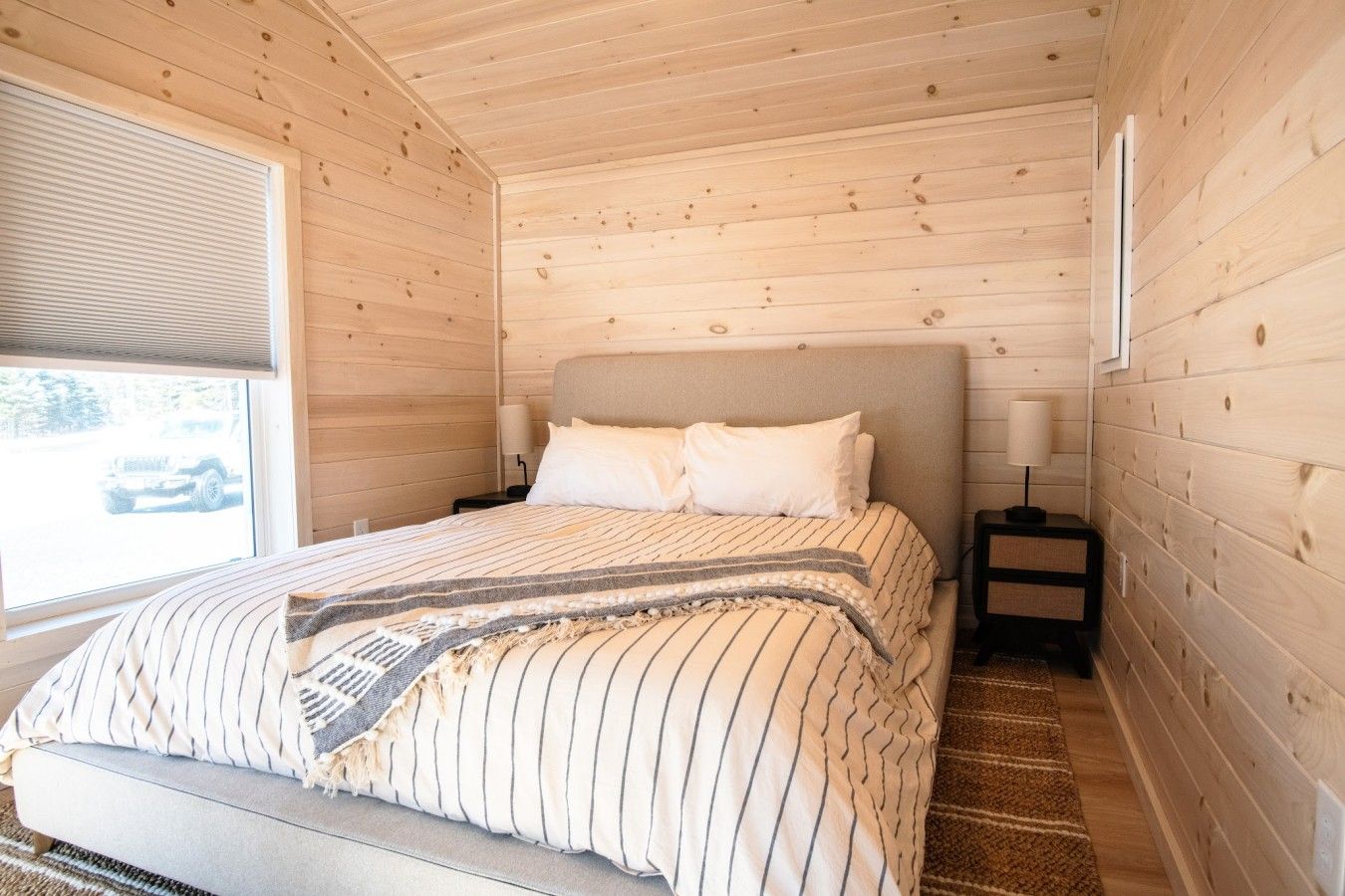 A bedroom with a bed, bedside tables, a window, and a view of the outside.