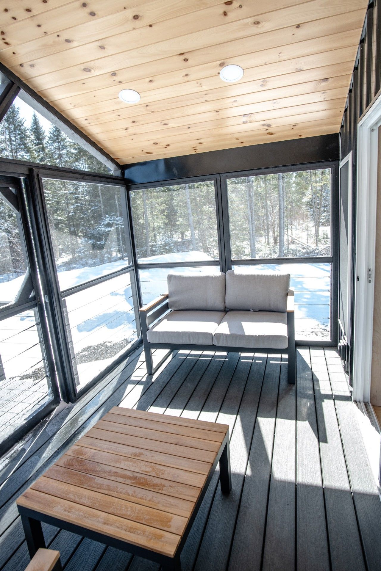 A covered outdoor seating area with a bench and a table, large windows, and a view of a snowy landscape.