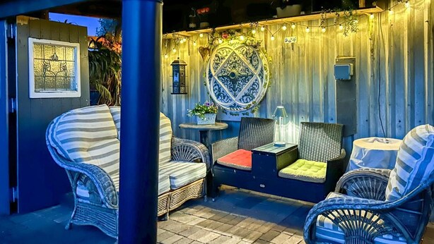 Un área de estar al aire libre con mobiliario de mimbre, un muro decorativo y luces colgantes.