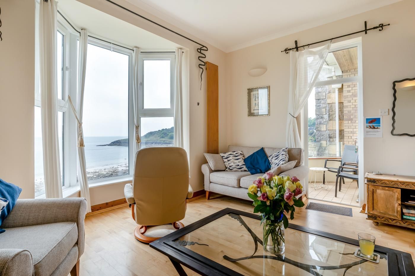 A living room with a sofa, armchairs, a glass coffee table, and a view of the sea through large windows.