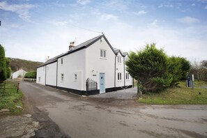 A white two-story house with a black roof, a blue door, and a small garden with a bush.
