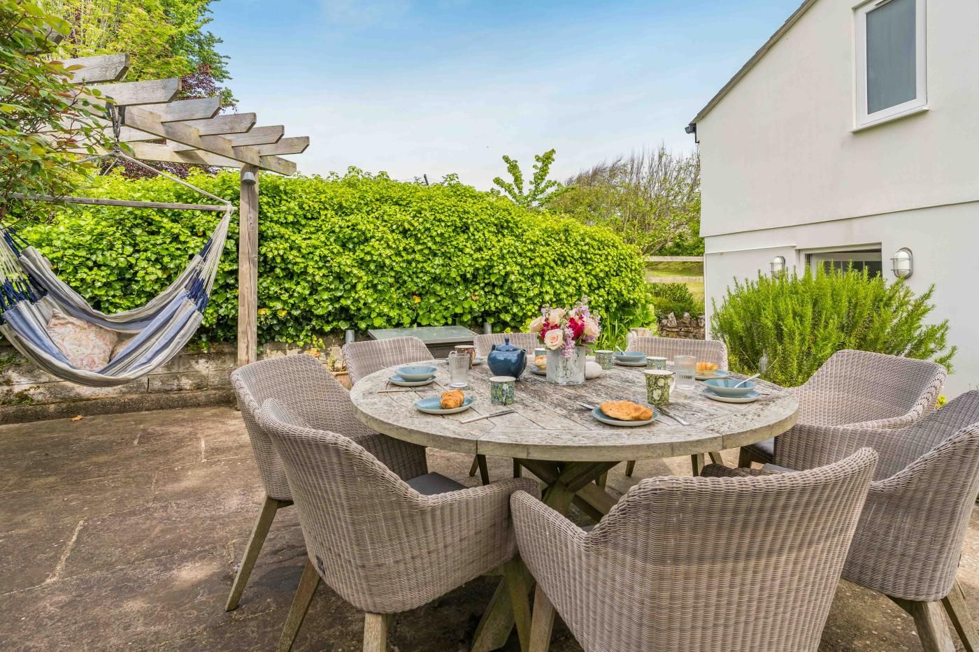 Une table de jardin ronde, entourée de chaises en osier, le tout agrémenté de verdure et adossé à un bâtiment blanc.