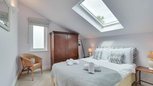 Ein Schlafzimmer mit Dachfenster, einem Bett mit Kissen, einem Holzschrank, einem Korbstuhl und einer Nachttischlampe.