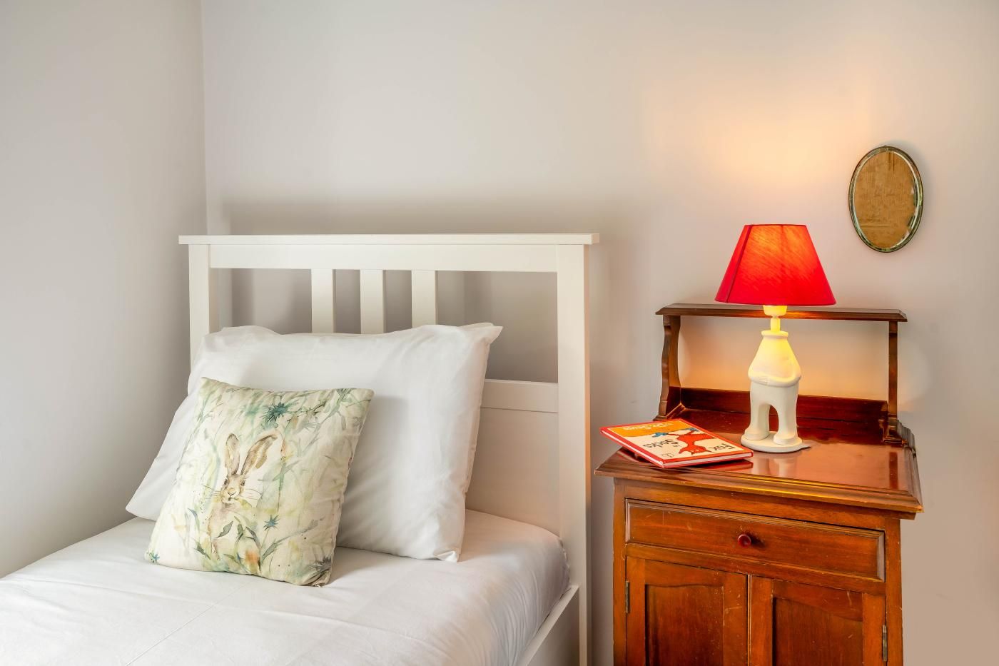 A bed with white bedding and a floral pillow, a wooden nightstand with a red lamp, and a small book.