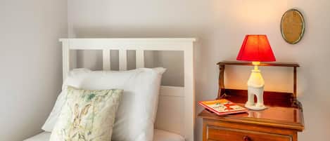 A bed with white bedding and a floral pillow, a wooden nightstand with a red lamp, and a small book.