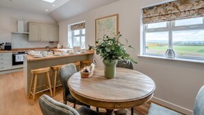 A modern kitchen with a central island, a dining area with a round table, and a view of a green landscape through large windows. - 2 Bed in Alyth (oc-e31854) (Auchrannie)