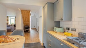 A modern kitchen with grey cabinets, a wooden countertop, and a toaster. A dining area with a brown sofa and a baby stroller is visible in the background. - 3 Bed in Nant Peris (oc-f31804) (Nant Peris)