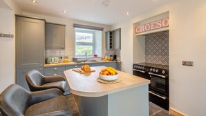 A modern kitchen with grey cabinets, a central island with fruit bowl, and a built-in oven. - 3 Bed in Nant Peris (oc-f31804) (Nant Peris)