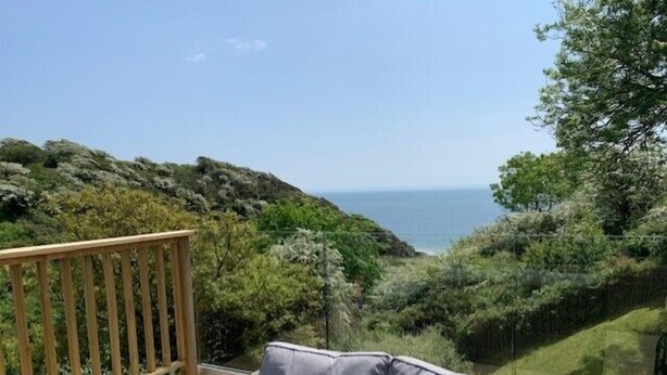 Un canapé en osier agrémenté de coussins, placé sur un balcon donnant sur un paysage côtier avec des arbres et la mer.