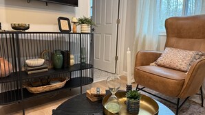A living room with a black coffee table, a brown leather armchair, a black metal shelf with decorative items, and a large window with a view of trees.