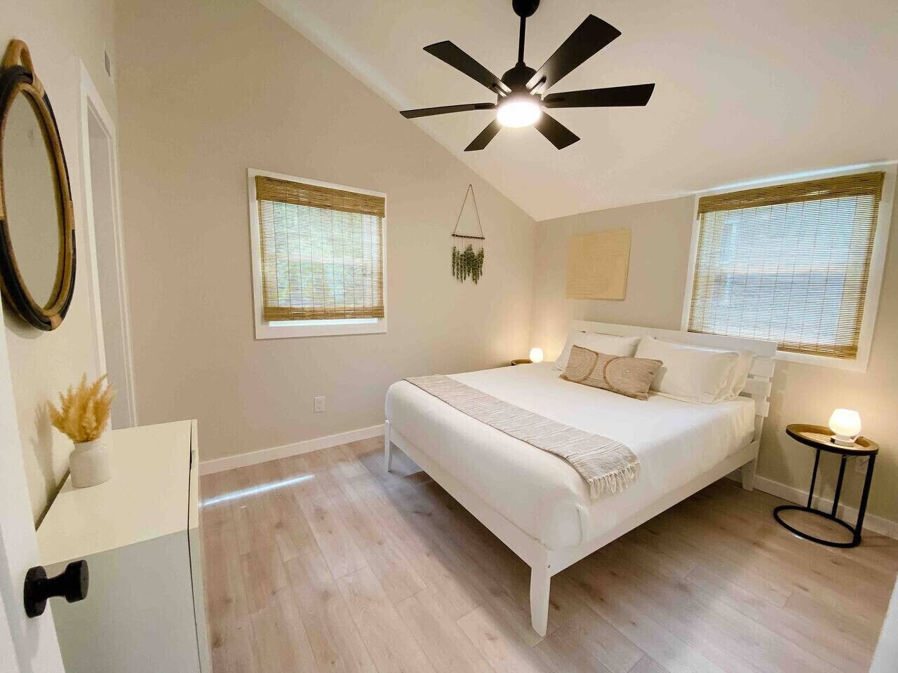 A bedroom with a bed, a ceiling fan, two windows with blinds, a bedside table with a lamp, and a wall-mounted mirror.