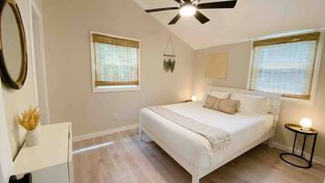 A bedroom with a bed, a ceiling fan, two windows with blinds, a bedside table with a lamp, and a wall-mounted mirror.