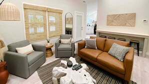 A modern living room with a grey sofa, a brown leather sofa, and a grey armchair. There is a coffee table with a white cloth and a remote control. The room has large windows with blinds, a fireplace, and a kitchen in the background. - Endless Summer Cottage - walk to the GOTL strip (Geneva)