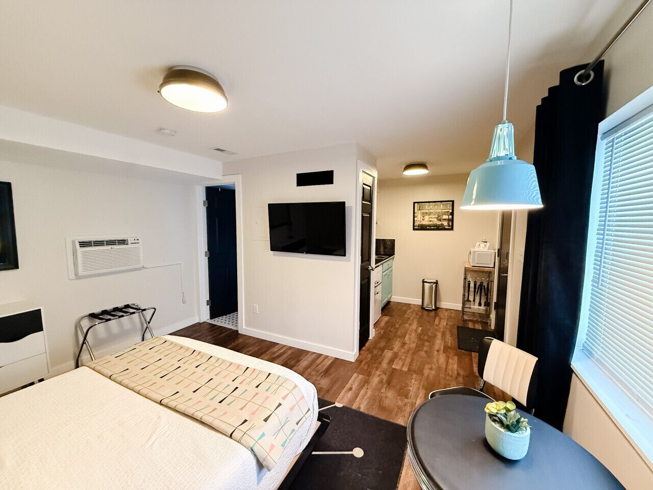 Ein kleines Hotelzimmer mit Bett, Tisch, Schreibtisch, Stuhl, einer Pendelleuchte und einem Fenster mit Jalousien.
