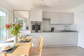 A modern kitchen with white cabinets, a built-in oven, and a sink. There is a dining table with chairs, a vase with flowers, and breakfast items. - 1 Bed in Gower (oc-hh9306) (Bishopston)