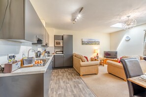 A modern kitchen with grey cabinets, a microwave, and a sink. There is a dining area with a sofa and a TV. - 3 Bed in Isel (oc-o32074) (Setmurthy)