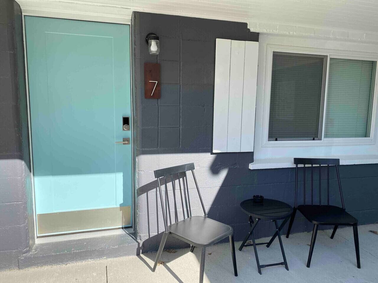 A teal door with a number '7' sign, two chairs, a small table, and a window with blinds.
