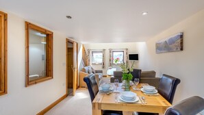 Een eetgedeelte met een houten tafel voor vier personen, omringd door lederen stoelen. Een televisie is aan de muur boven de bank in de achtergrond bevestigd.