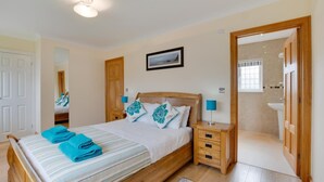 A bedroom with a wooden bed, a nightstand with a lamp, a bathroom visible through an open door, and a mirror on the wall. - 3 Bed in Gower (oc-hh23) (Port Eynon)