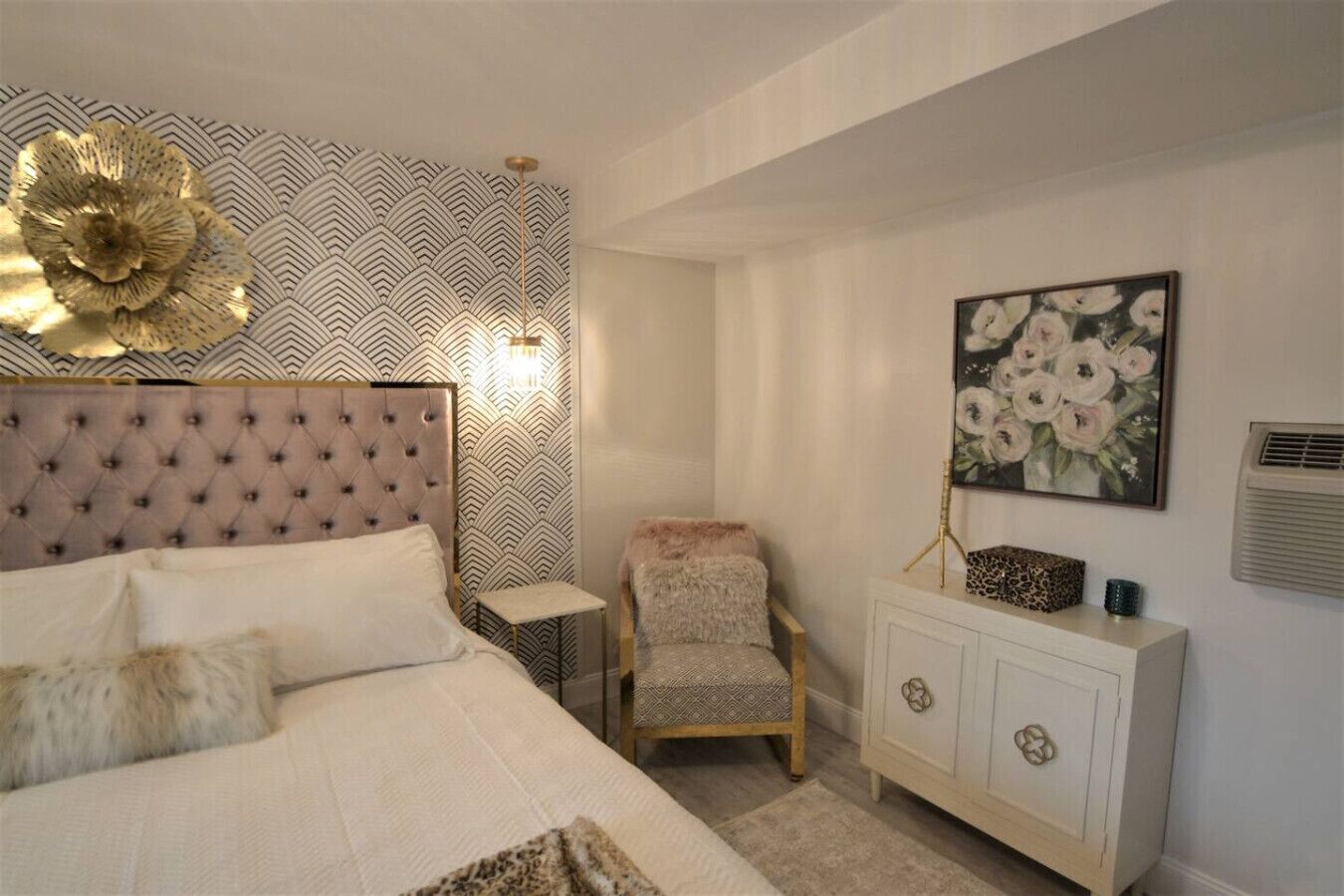 A bedroom with a tufted headboard, a patterned wallpaper, a floral painting, a leopard print side table, and a white cabinet.