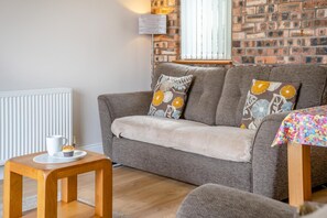 A cozy living room with a grey sofa, a wooden side table with a white mug, and a colorful tablecloth. - 2 Bed in Gower (oc-hh487) (Pen-clawdd)