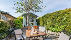 Ein Essbereich im Freien mit einem Tisch für zwei Personen, umgeben von Stühlen, einem Baum und einem Gebäude im Hintergrund.