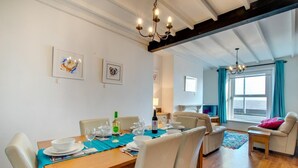 Uma sala de jantar com mesa de madeira, cadeiras brancas e um candelabro. Há quadros nas paredes, um sofá e uma janela com cortinas.
