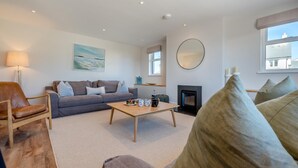A living room with a sofa, a coffee table, a fireplace, and a large window. - 4 Bed in St. Merryn (oc-f31095) (Saint Merryn)