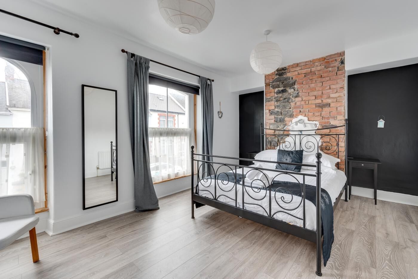A modern bedroom with a metal bed frame, a brick accent wall, a large window with curtains, and a white ceiling light.