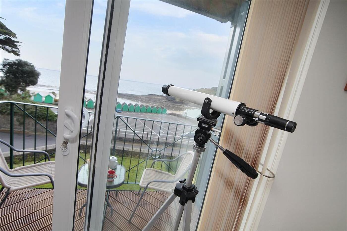 A telescope mounted on a balcony railing, overlooking a coastal area with houses and a road.