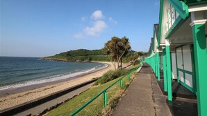 A coastal walkway with a beach, a green building with white trim, and a hill in the background. - 1 Bed in Langland (oc-hh147) (Newton)
