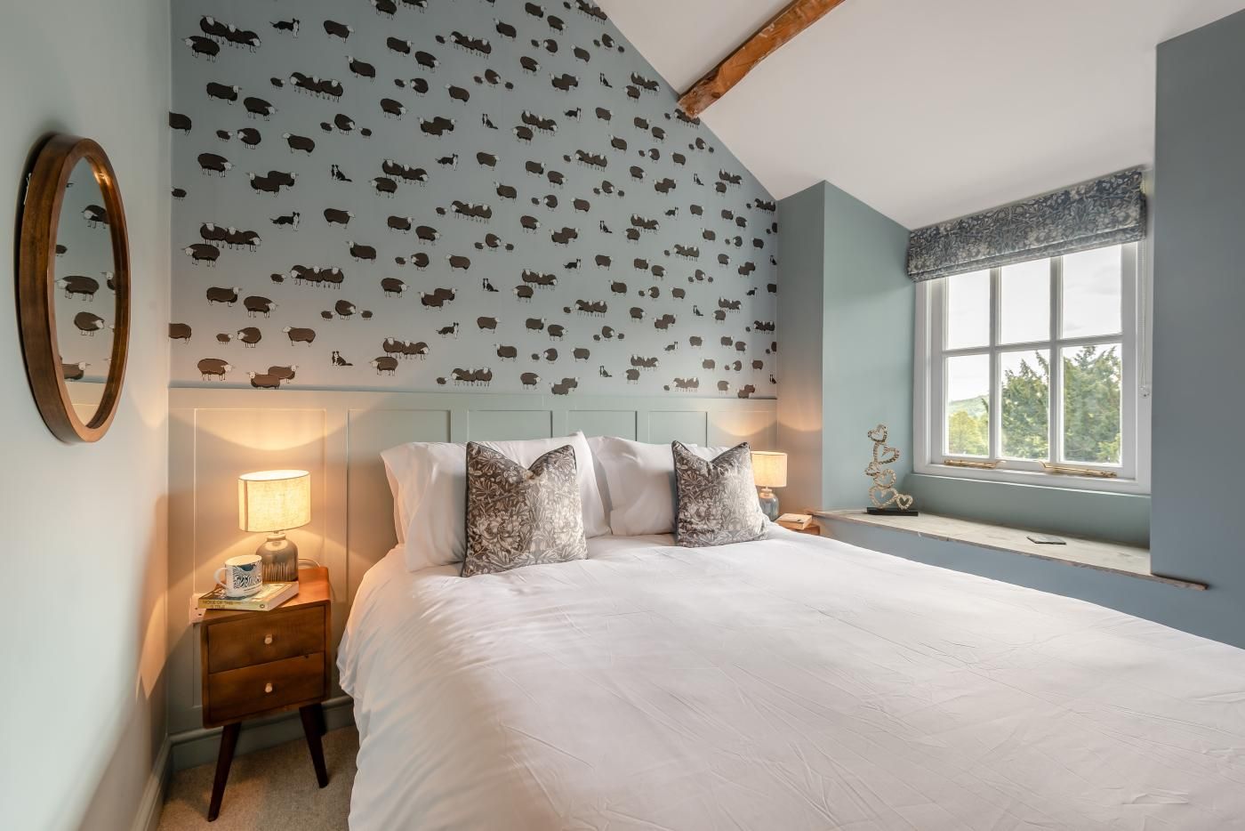 A bedroom with a large bed, a nightstand with a lamp, a mirror, and a window with a view.