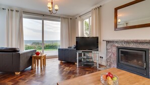 Een ruime woonkamer met een open haard, een televisie en een eethoek met fruit op tafel.