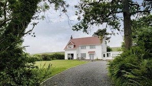 Een modern huis met een oprit van grind dat leidt naar een witte gevel, omringd door weelderige begroeiing en bomen.