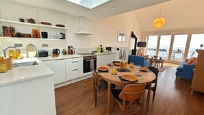 A modern kitchen with white cabinets, a dining area with wooden chairs, and a view of the sea through large windows. - 2 Bed in Craster (oc-c25099) (Dunstan)