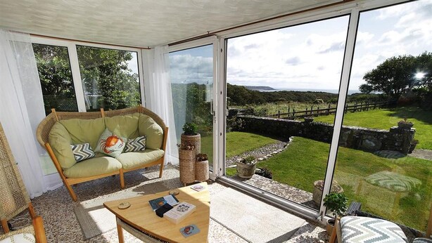 A room with a large sliding glass door leading to a grassy outdoor area, a wicker sofa, a wooden table, and a wicker chair. - 3 Bed in Overton (oc-hh2103) (Port Eynon)