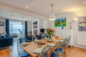 A dining area with a wooden table set for four, surrounded by chairs, with a view of the sea through large windows. - 3 Bed in Craster (oc-c25115) (Dunstan)
